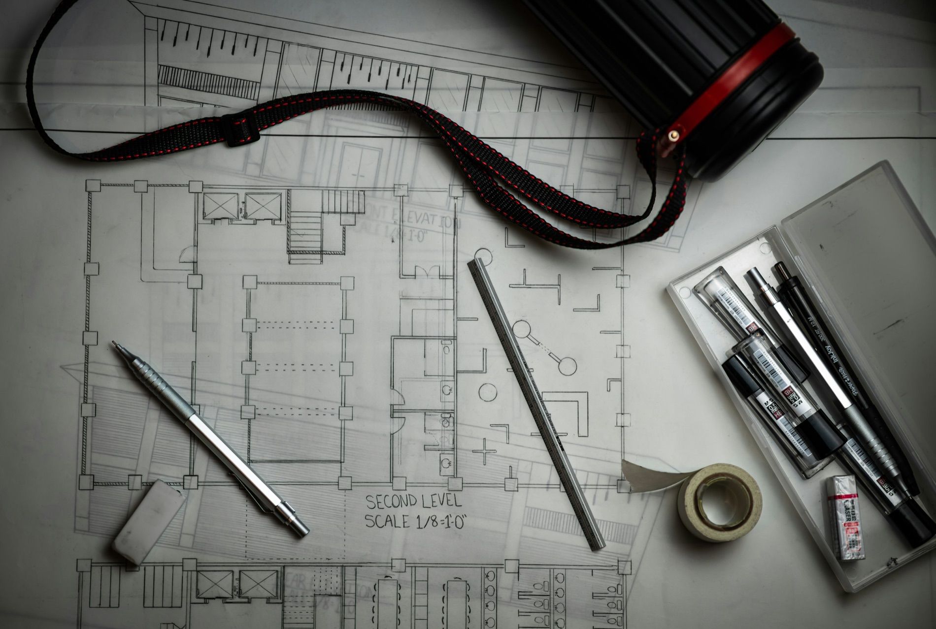 A floor plan on a drafting table with technical pens, a scale rule and a rolled drawing tube