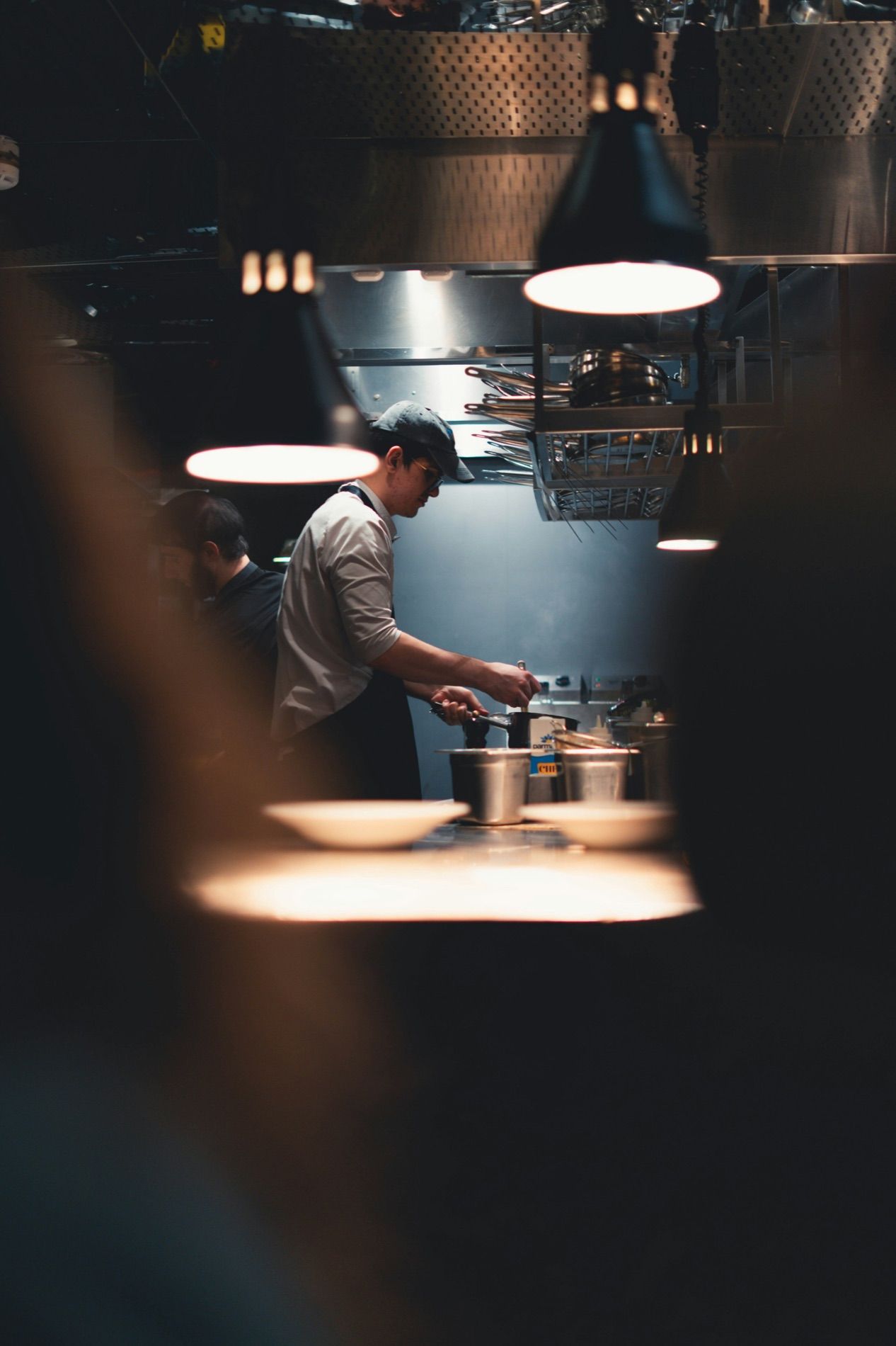 A cook at the stove seen through the gap of a service pass in a dark kitchen